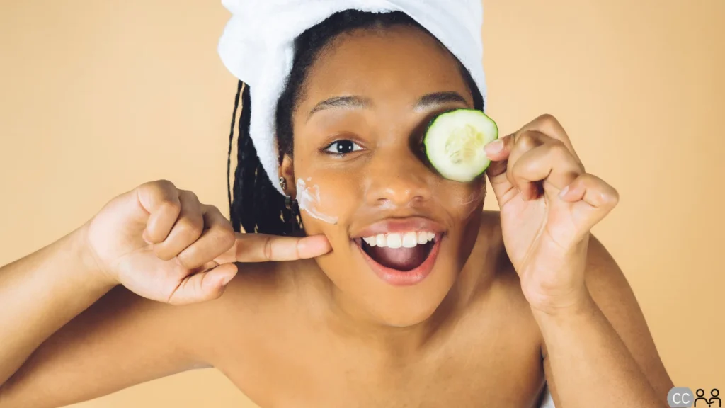 A woman with a cucumber over her eye pointing at lotion on her check